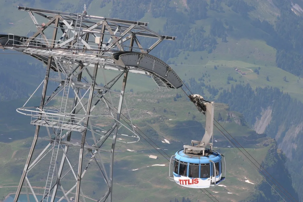 Eine Seilbahn, gekennzeichnet mit 'Titlis', fährt über eine grüne alpine Landschaft, unterstützt von einem großen Metallturm.