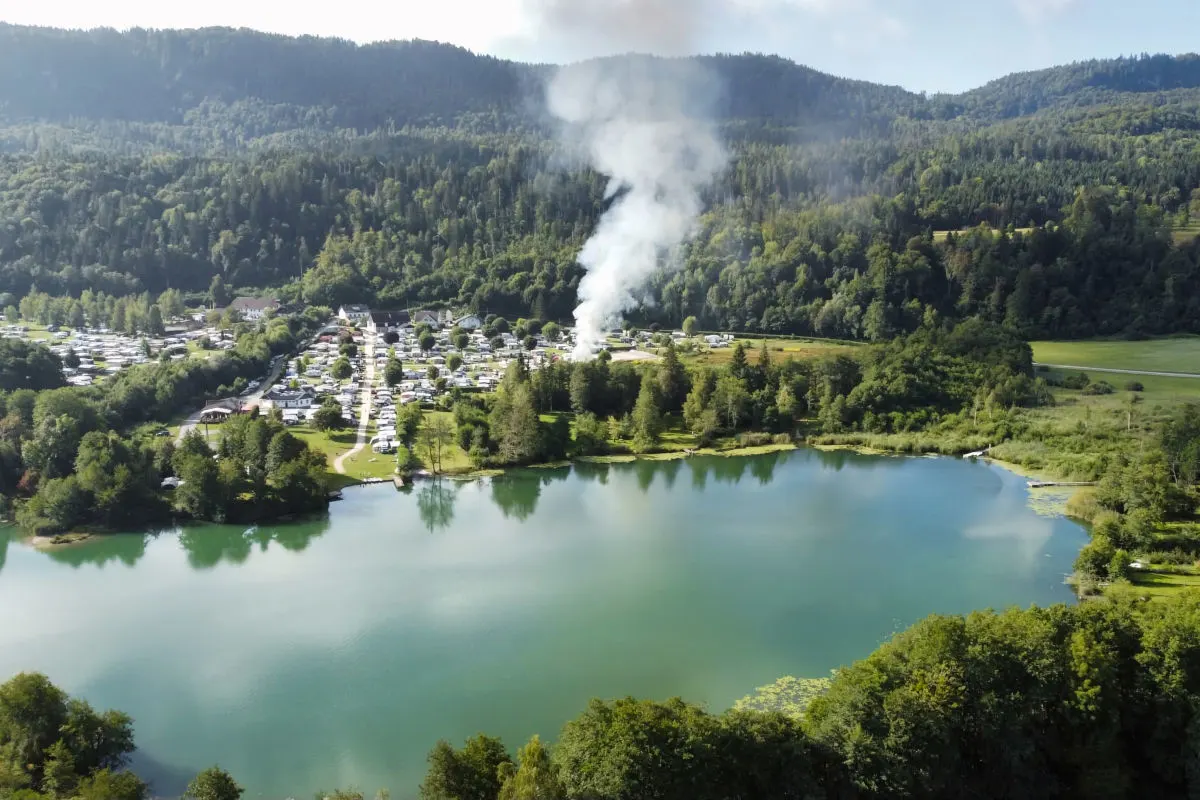 Eine Rauchwolke erhebt sich über einem Campingplatz, der von Wald und einem See umgeben ist. Die Aufnahme zeigt die Szenerie aus der Vogelperspektive.