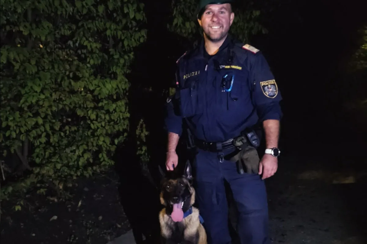 Ein Polizist steht nachts im Freien neben einem Hund. Im Hintergrund sind Büsche sichtbar.