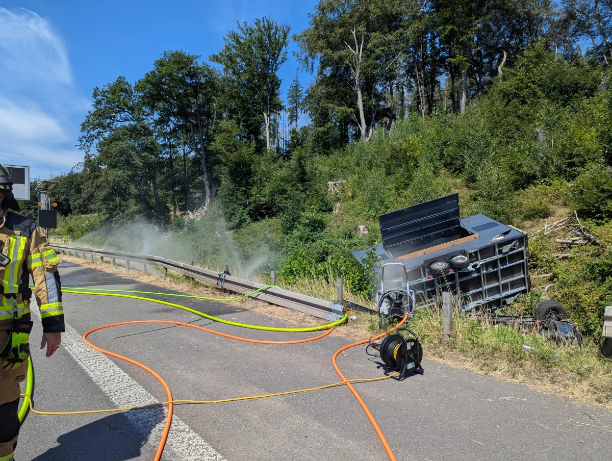 Ein umgekippter Anhänger liegt an einer Böschung neben einer Straße. Ein Feuerwehrmann ist vorne links im Bild.