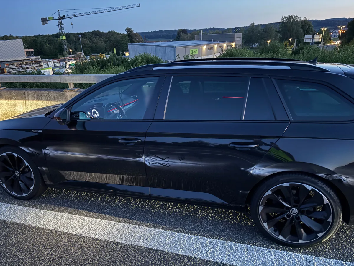 Ein schwarzer Kombi mit sichtbarer Delle an der Fahrerseite steht auf einer Brücke bei Abenddämmerung. Im Hintergrund sind Baukräne und Bäume zu sehen.