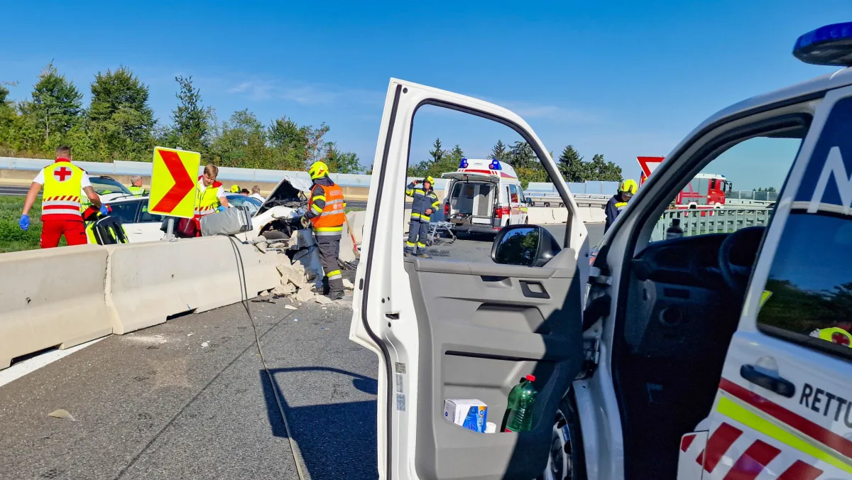 Ein weißes Auto ist in eine Autobahnleitplanke geprallt. Rettungskräfte und Feuerwehrleute arbeiten an der Unfallstelle, während Einsatzfahrzeuge den Bereich sichern.