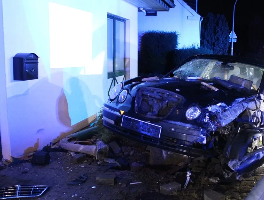 Ein schwarzes Auto steht mit stark beschädigter Front in der Hauswand. Trümmer liegen verstreut um das Fahrzeug.
