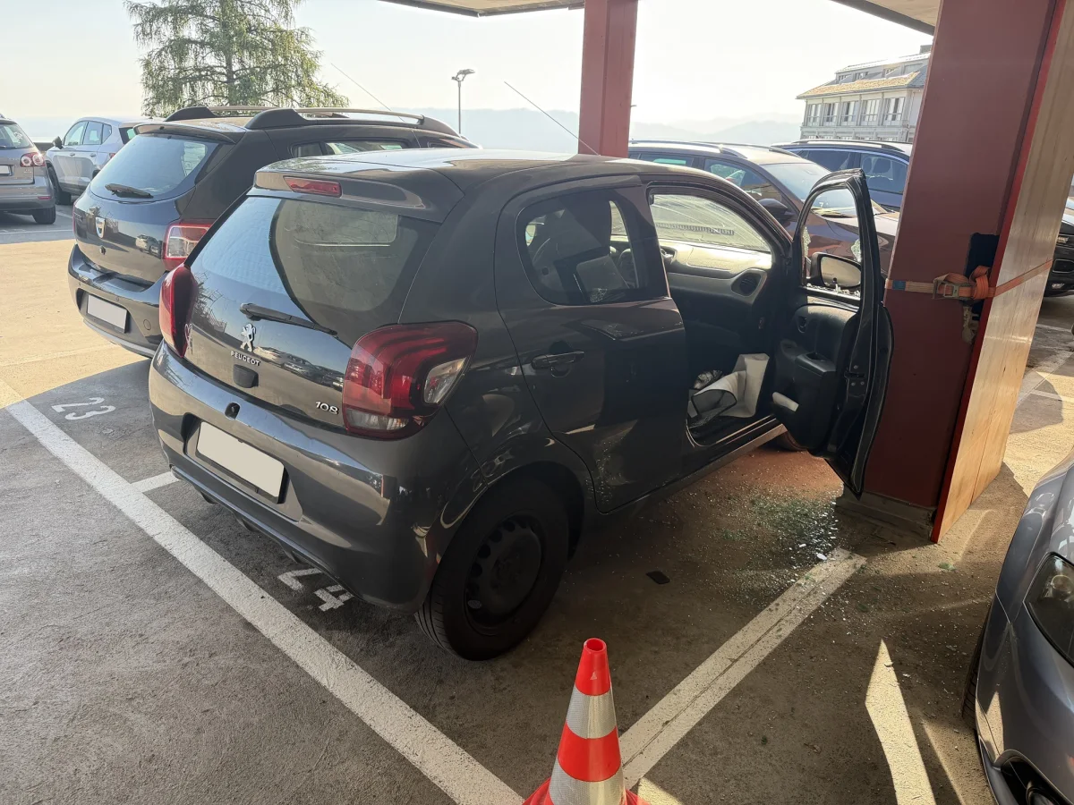 Das Foto zeigt einen grauen Peugeot 108 mit geöffneter Fahrertür in einem Parkdeck. Neben dem Fahrzeug liegt zerbrochenes Glas auf dem Boden.
