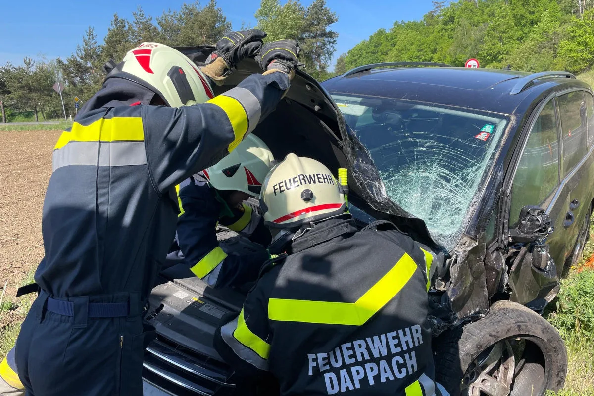 Mehrere Feuerwehrleute in Schutzkleidung arbeiten an einem stark beschädigten schwarzen Auto am Straßenrand. Die Windschutzscheibe des Autos ist zerstört, und die Feuerwehrleute scheinen Rettungsarbeiten durchzuführen.