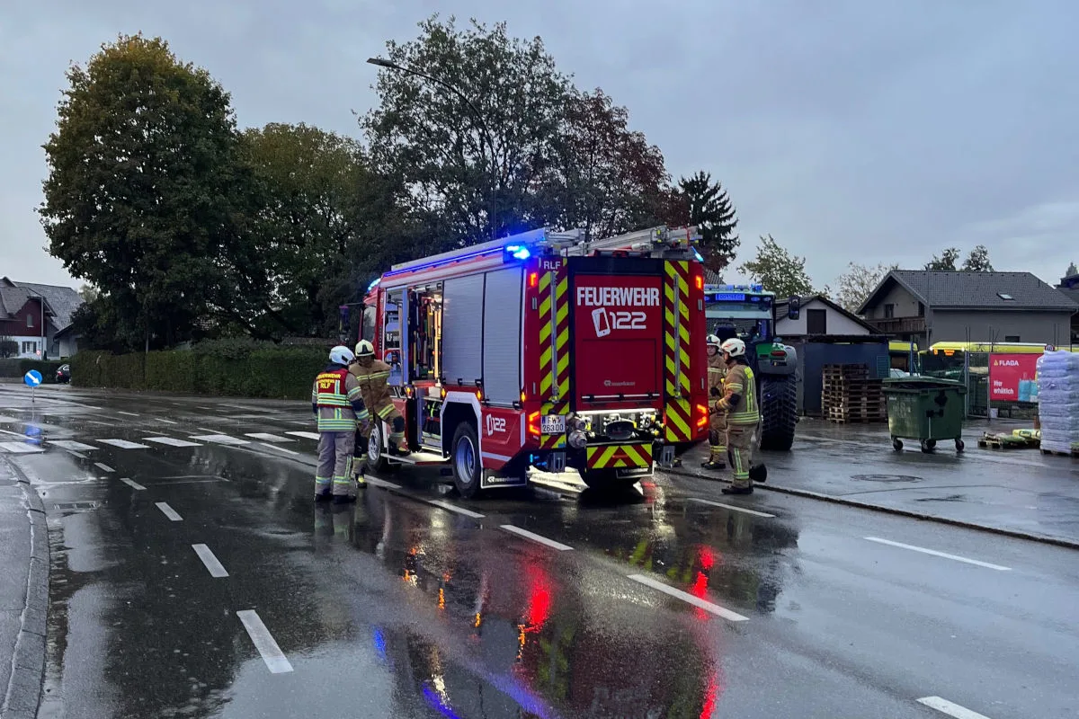 Feuerwehrleute stehen neben einem rot-weißen Einsatzfahrzeug auf einer regennassen Straße. Der Himmel ist bewölkt.