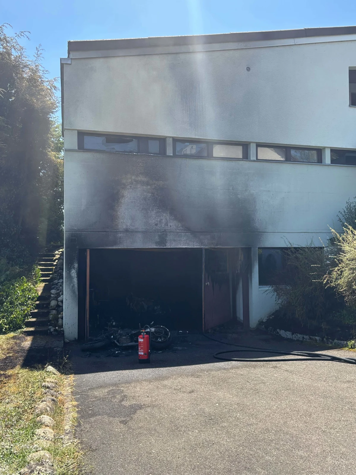 Eine Garage mit Brandschäden, in der ein verbranntes Motorrad steht. Ein Feuerlöscher befindet sich davor auf der Auffahrt.