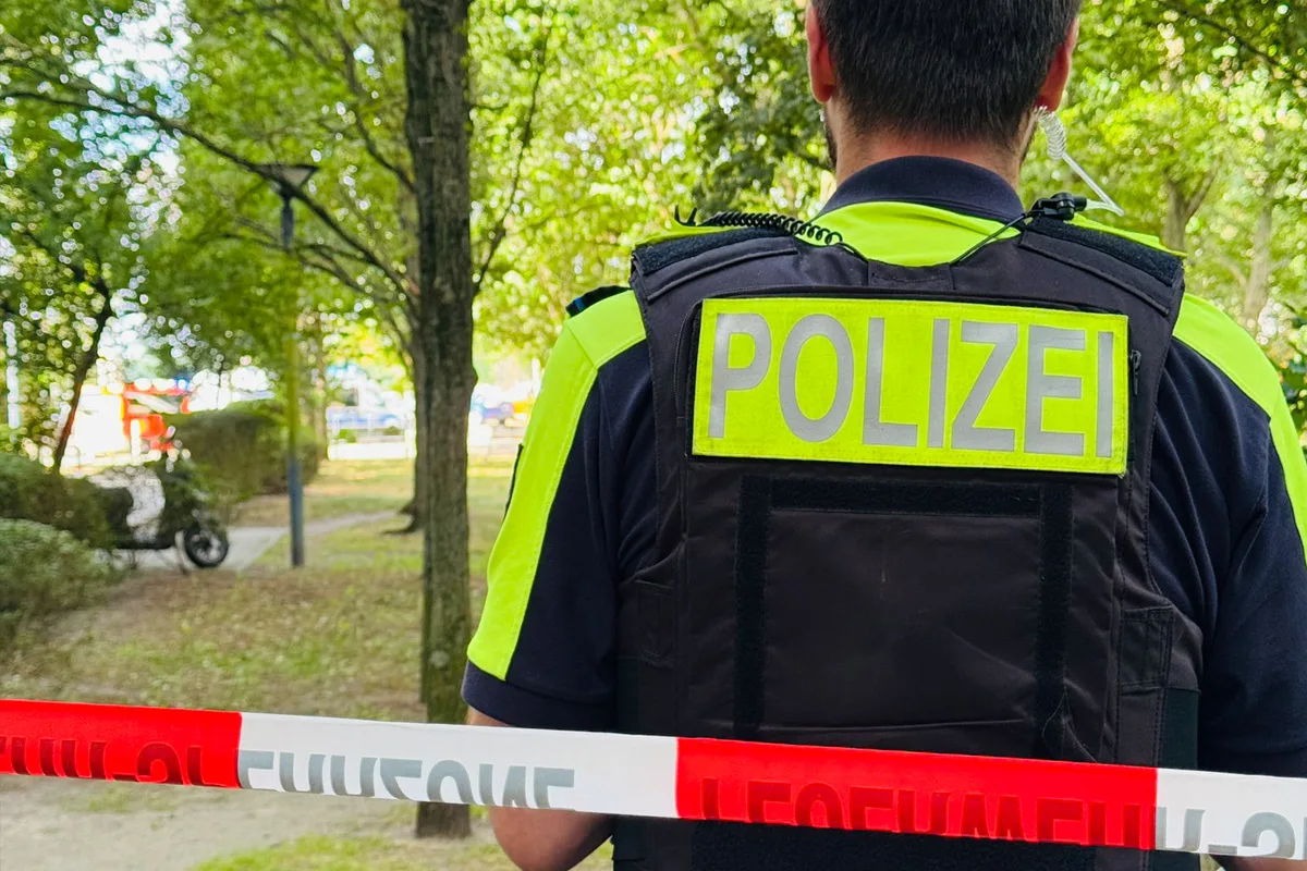 Ein Polizist in gelb-schwarzer Uniform steht mit dem Rücken zur Kamera hinter einem rot-weißen Absperrband in einem parkähnlichen Areal.
