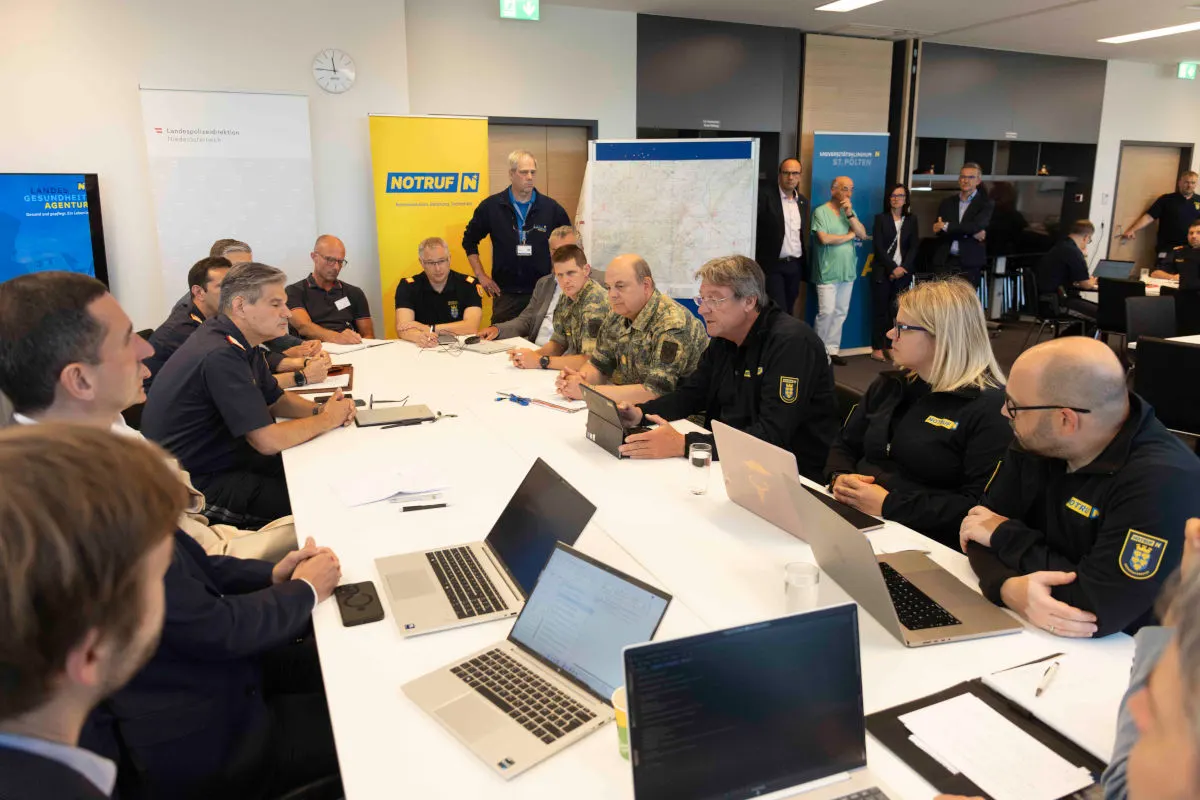 Mehrere Personen sitzen in einem Besprechungsraum an einem Tisch mit Laptops. Einige tragen Uniformjacken mit Aufnähern, während andere in ziviler Kleidung sind.