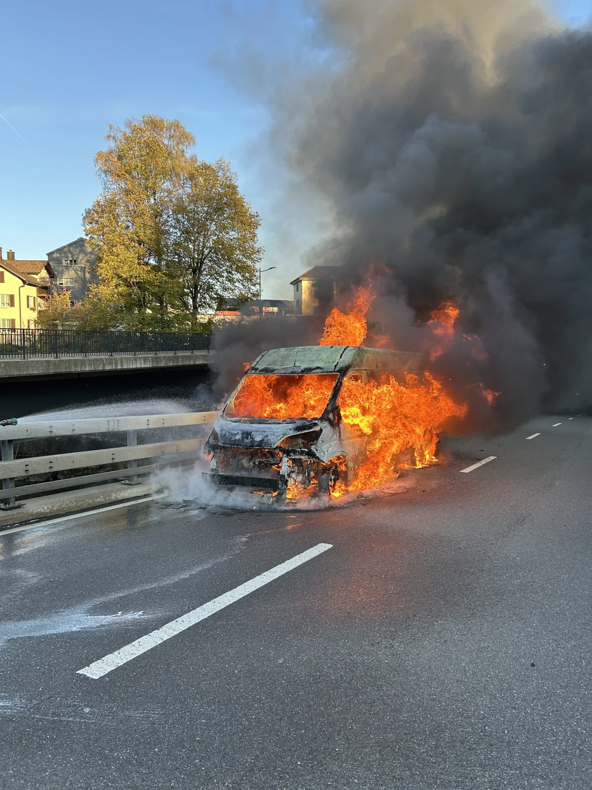 Ein Fahrzeug steht mitten auf der Straße in Flammen, dichter Rauch steigt auf. Im Hintergrund sind Gebäude und Bäume sichtbar.