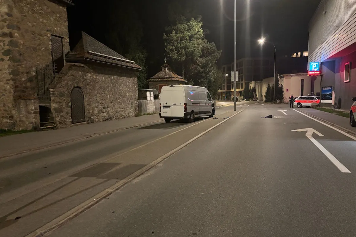 Bei Nacht steht ein weißer Lieferwagen auf der Straße, im Hintergrund ist ein Polizeiauto zu sehen. Die Szenerie wirkt ruhig, und es sind keine Menschen auf der Straße sichtbar.