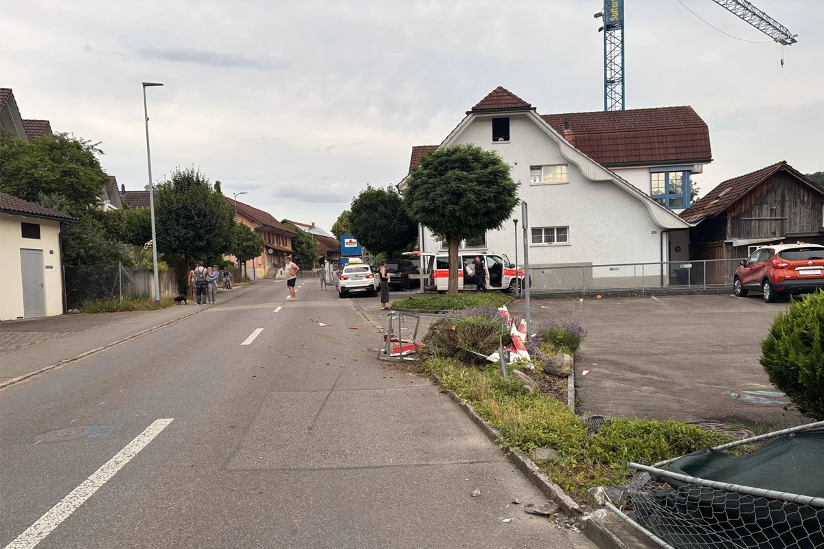 Ein Polizeifahrzeug parkt vor einem weißen Haus an einer Dorfstraße, umgeben von Menschen, mit einem beschädigten Zaun sichtbar.