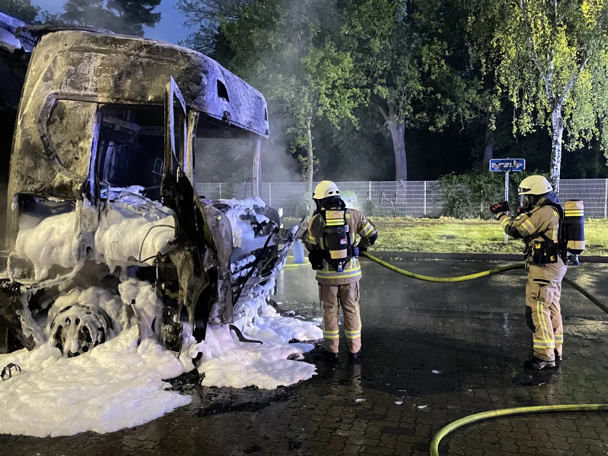 Zwei Feuerwehrleute mit Atemschutzgeräten sprühen Schaum auf einen ausgebrannten LKW bei Nacht. Im Hintergrund sind Bäume und ein Zaun.