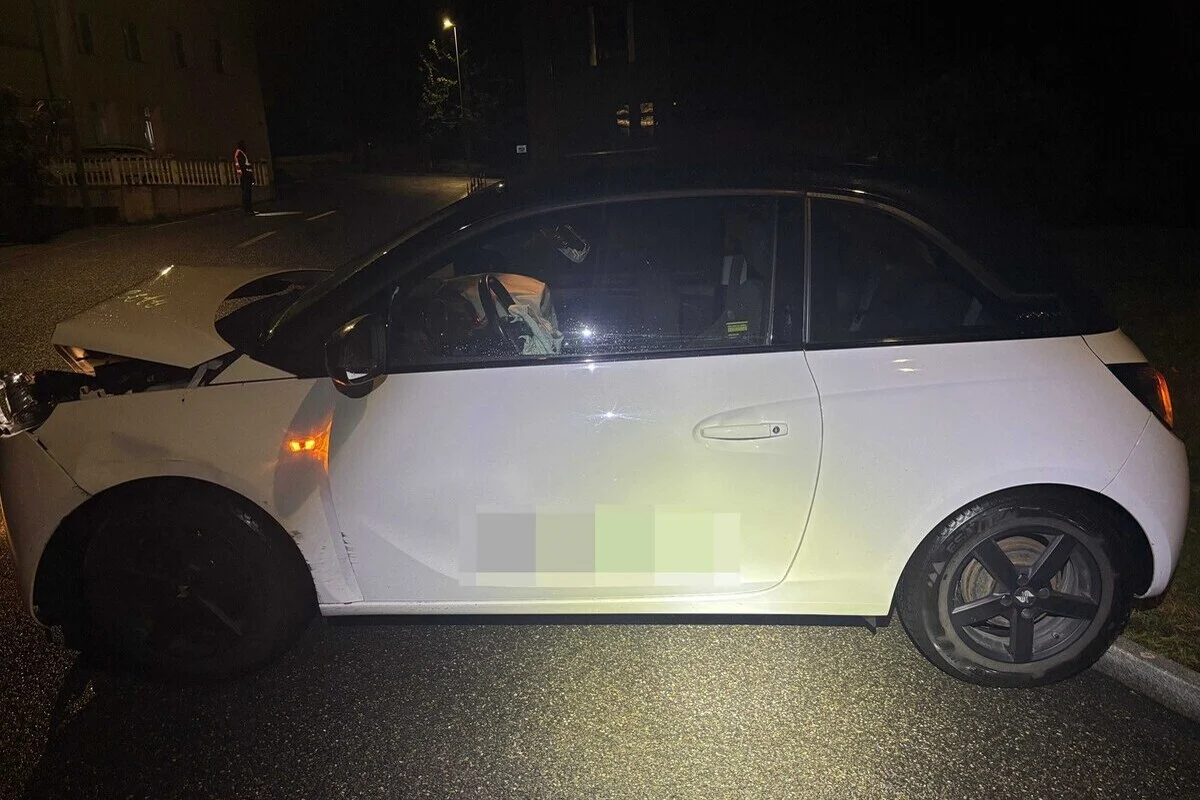 Ein weißes Auto mit geöffneter und beschädigter Motorhaube steht bei Nacht auf einer nassen Straße. Im Hintergrund ist eine Straßenlaterne sichtbar.