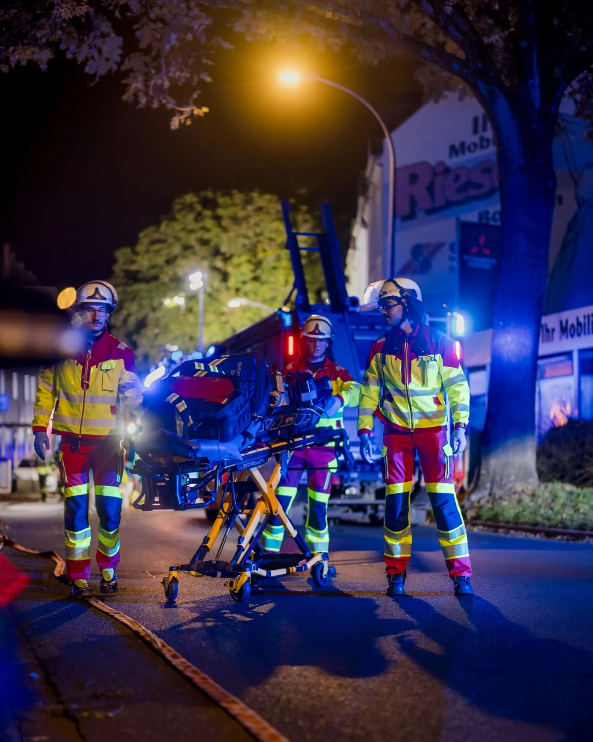 Drei Rettungskräfte stehen auf einer nächtlichen Straße neben einer Trage. Im Hintergrund sind Einsatzfahrzeuge beleuchtet.
