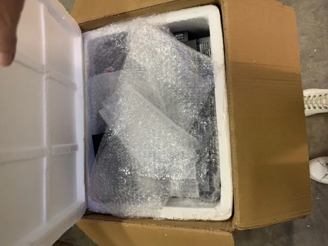 Ein brauner Karton mit geöffnetem Deckel, darin Verpackungsmaterial wie Luftpolsterfolie zu sehen.