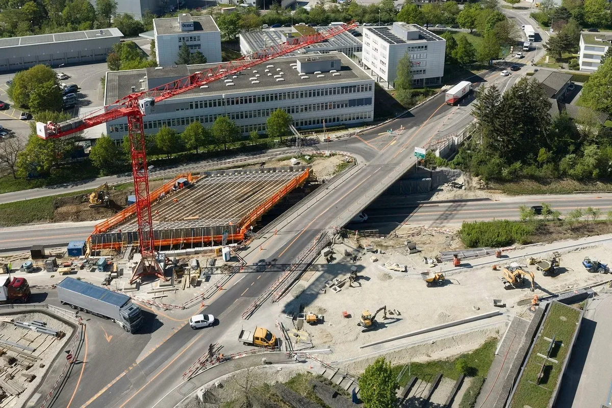 Die Aufnahme zeigt eine große Brückenbaustelle mit einem roten Kran, mehreren Baustellenfahrzeugen und umgeleitetem Verkehr. Im Hintergrund sind Bürogebäude und Straßen zu sehen.