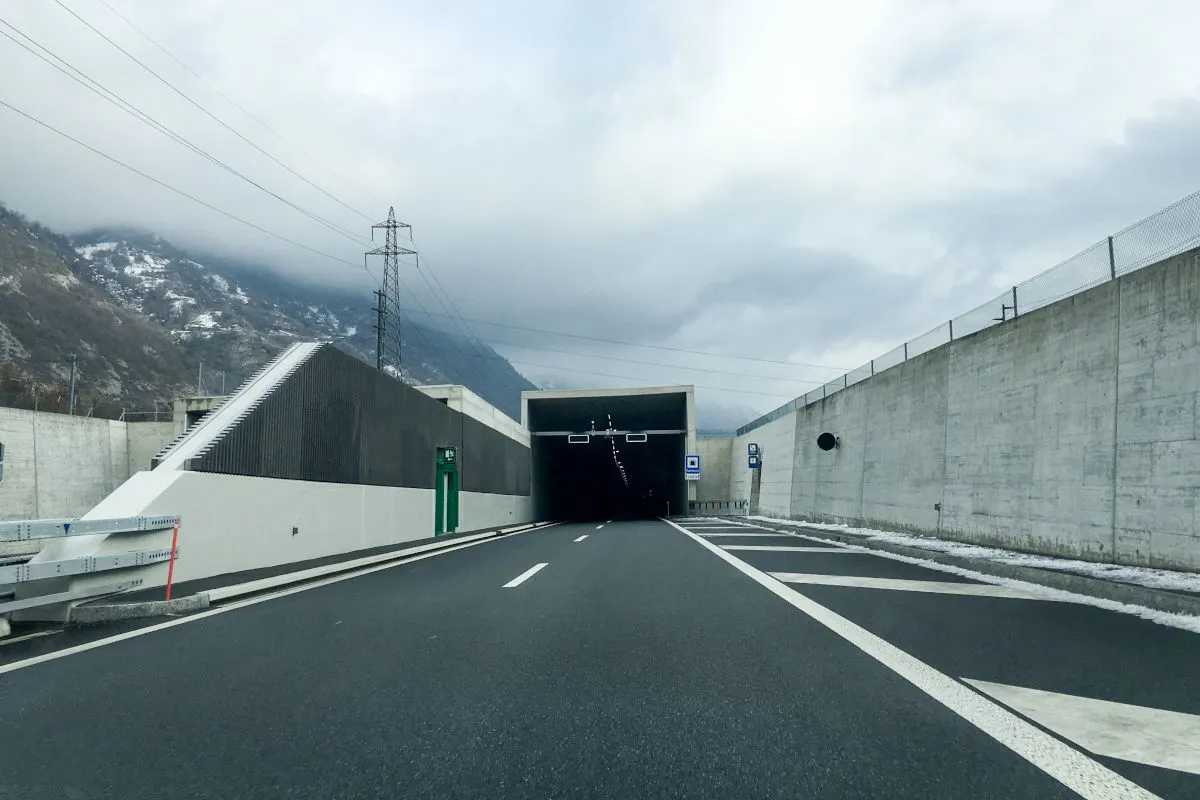 Eine Fahrbahn führt geradeaus in einen großen Straßentunnel. Die Umgebung ist bergig und der Himmel stark bewölkt.