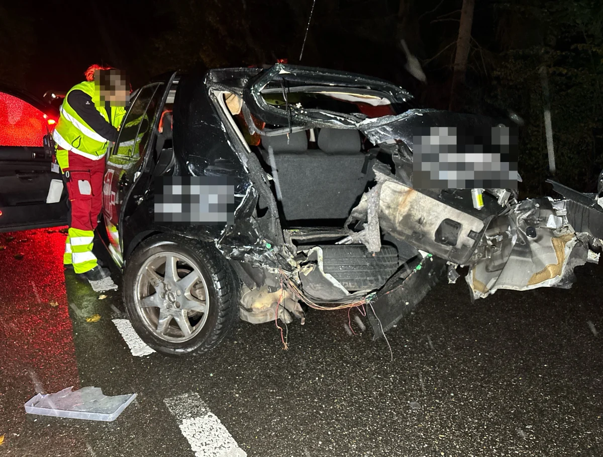 Ein Auto ist stark beschädigt mit einem großen Schaden am Heck. Daneben steht ein Rettungsmitarbeiter in reflektierender Kleidung.
