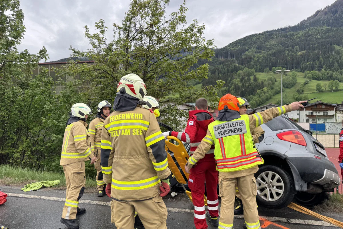 Mehrere Feuerwehrleute und ein Sanitäter stehen um ein graues Auto, das auf dem Straßenrand verunglückt ist. Im Hintergrund sind Berge und Bäume zu sehen.