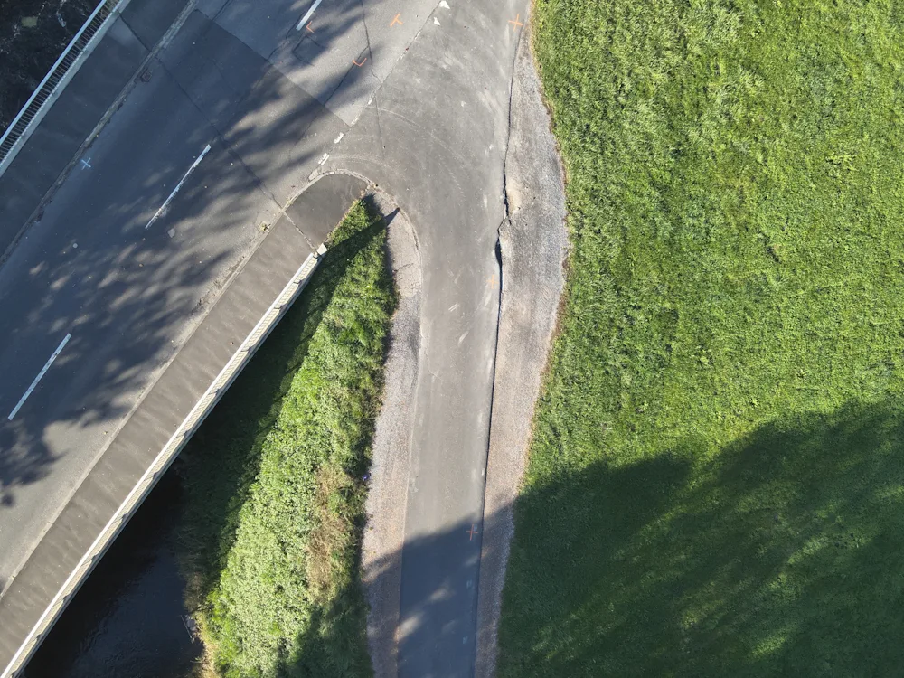 Luftaufnahme einer Straßenkurve mit einem großen Riss, der die Fahrbahn erheblich beschädigt. Die Umgebung besteht aus Gras und Asphalt.
