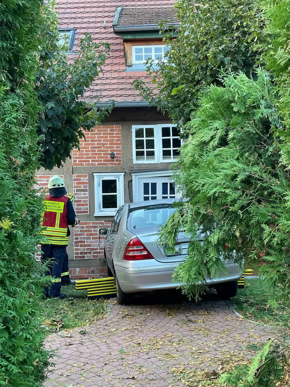 Ein silbernes Auto steht unmittelbar vor einer Hauswand, flankiert von grünen Bäumen, während ein Einsatzkraft daneben steht.