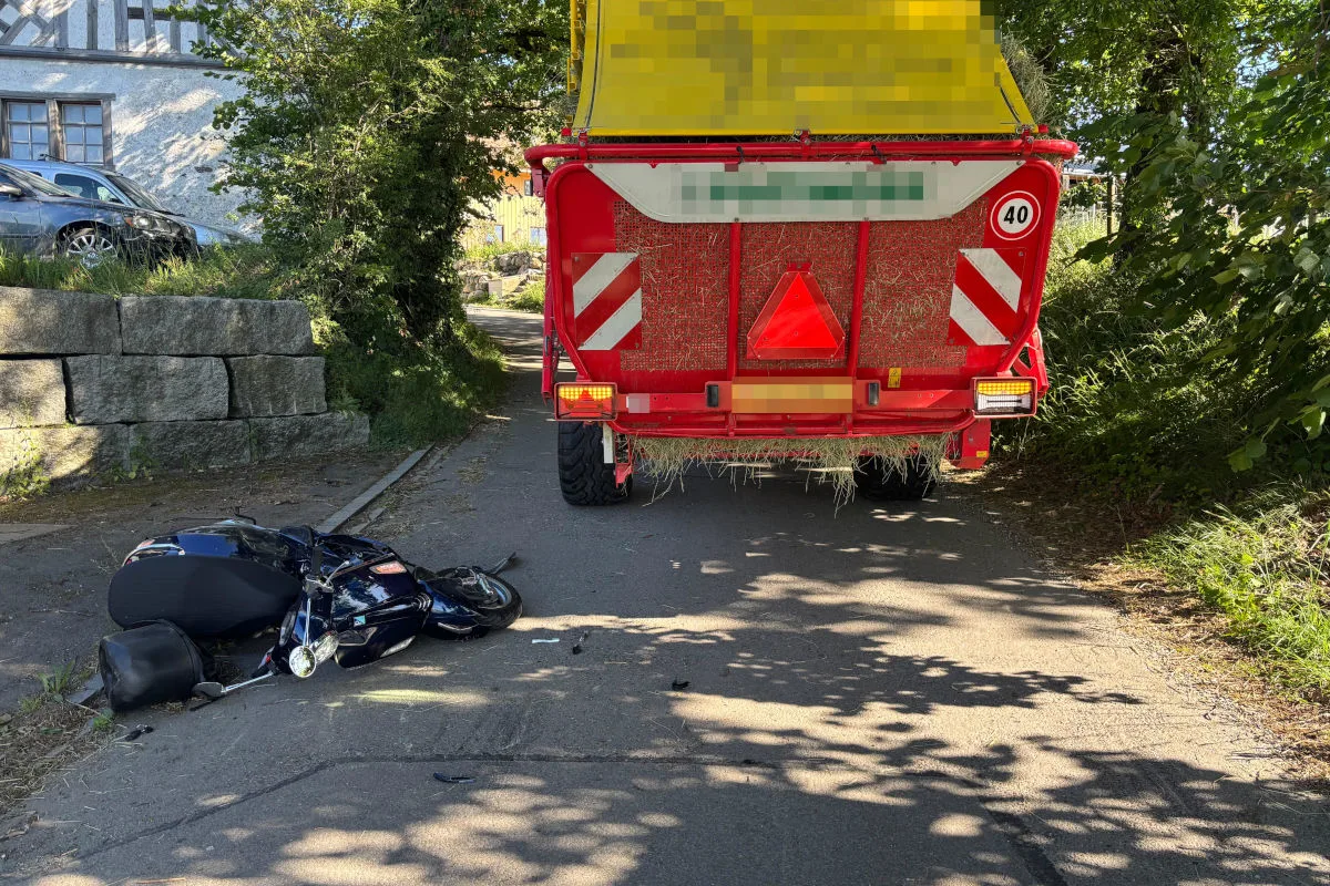 Ein gestürztes Motorrad liegt neben einem landwirtschaftlichen Anhänger auf einer schmalen, von Bäumen gesäumten Straße. Im Hintergrund sind ein Fachwerkhaus und eine Steinmauer zu sehen.