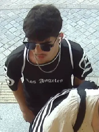 Ein Mann trägt eine Sonnenbrille, ein schwarzes T-Shirt mit Aufdruck und schaut leicht nach unten. Die Umgebung ist gepflastert.