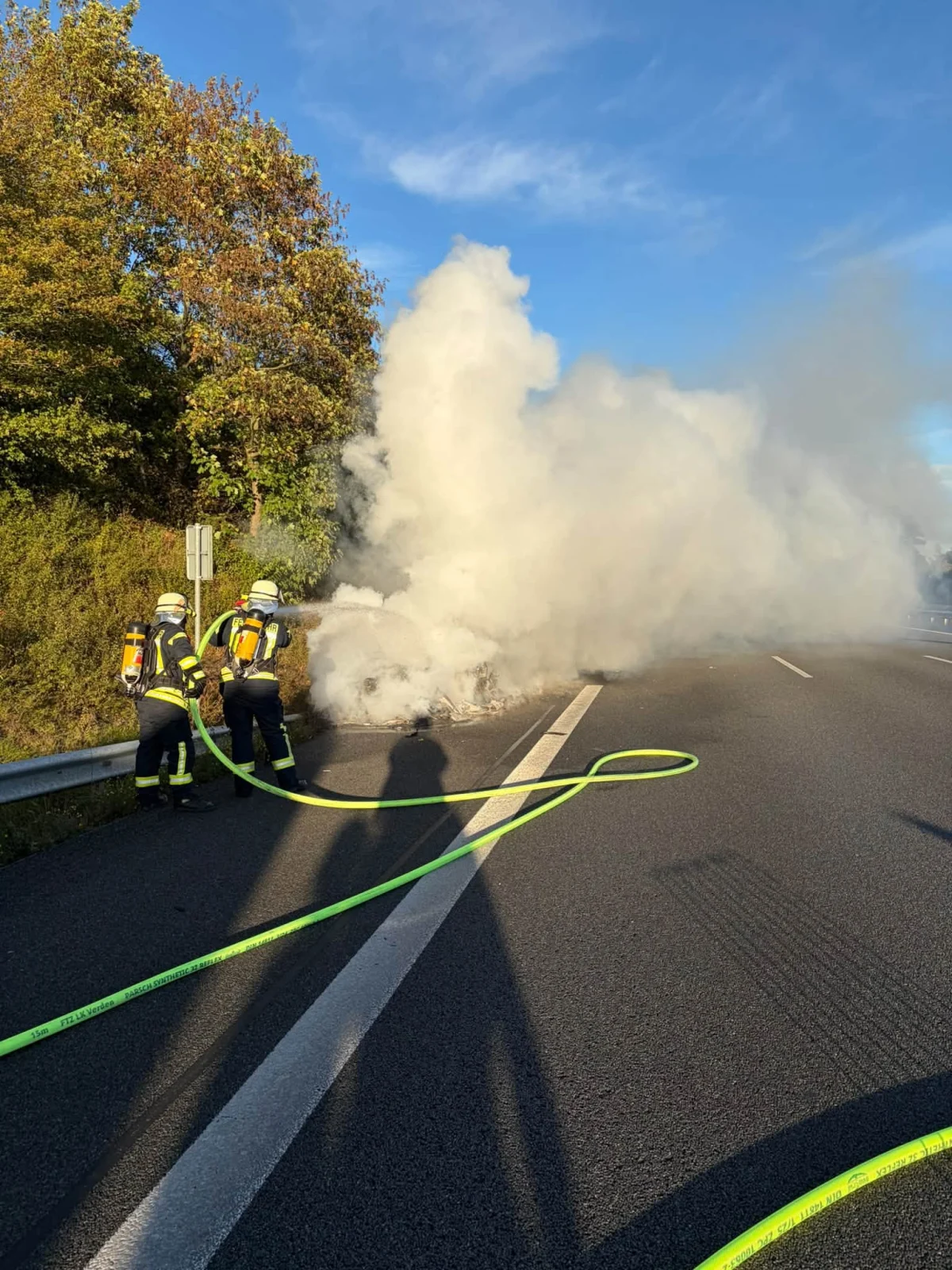 Zwei Feuerwehrmänner bekämpfen einen Brand auf einer Autobahn. Dichte Rauchwolken steigen auf und verdecken die Sicht.