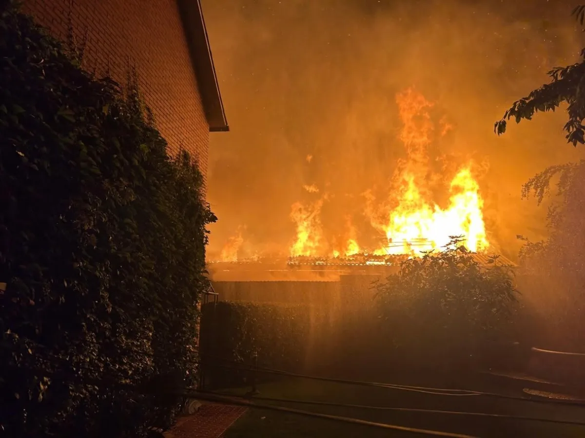 Ein großes Feuer lodert hinter einem Gebäude in der Nacht, dichte Rauchschwaden steigen auf. Pflanzen und ein Ziegelgebäude sind im Vordergrund sichtbar.