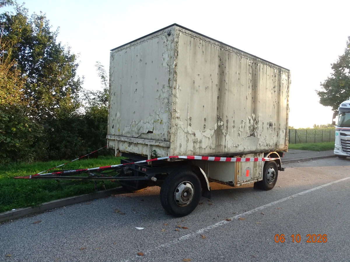 Ein verlassener, veralteter Anhänger steht am Straßenrand und ist mit rot-weißem Flatterband abgesperrt. Ein Lkw ist im Hintergrund sichtbar.