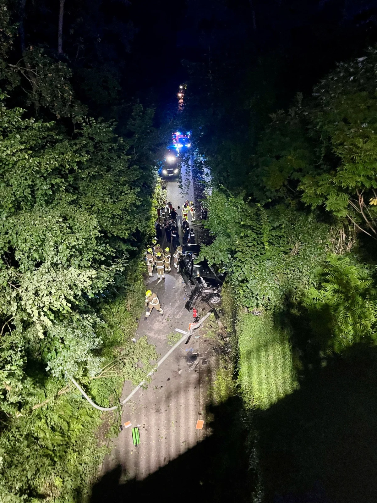 Bei Nacht zeigt das Bild eine Unfallstelle in einem Wald mit mehreren Feuerwehrleuten und einem beschädigten Fahrzeug auf einer engen Straße.