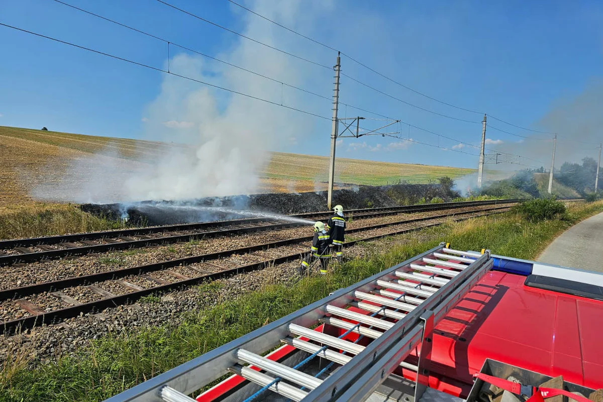 Zwei Feuerwehrleute spritzen Wasser auf einen Flächenbrand neben Bahngleisen. Im Hintergrund sind Felder zu sehen und Rauch steigt auf.