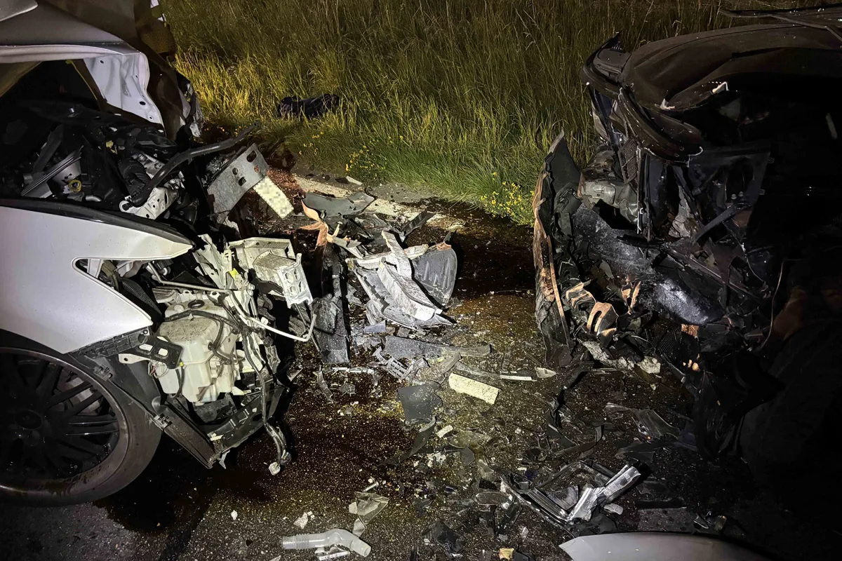 Ein schwerer Frontalzusammenstoß wird zwischen zwei Fahrzeugen gezeigt, die stark beschädigt sind. Trümmer liegen auf der Straße, umgeben von Grasflächen.