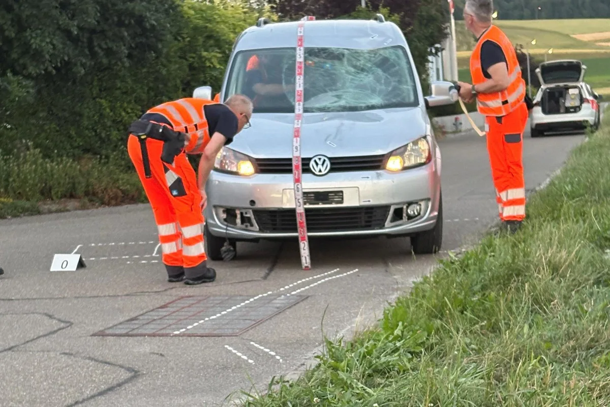 Zwei Ermittler in orangenen Westen inspizieren einen beschädigten silbernen VW auf einer ländlichen Straße am Abend.