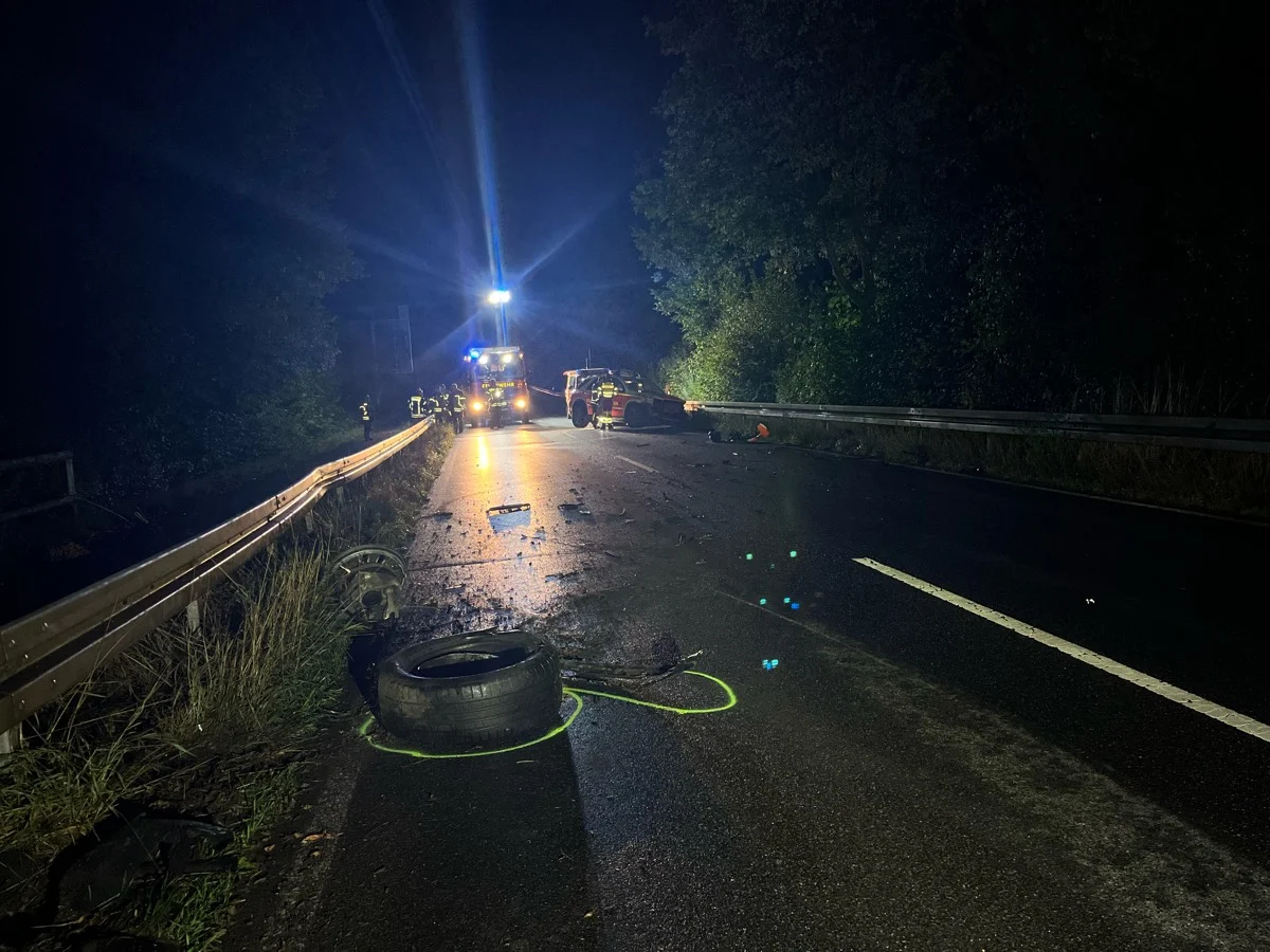 Auf einer dunklen Landstraße sind Einsatzkräfte bei einem Unfall im Einsatz. Trümmer und ein Fahrzeug mit Blaulicht sind sichtbar.