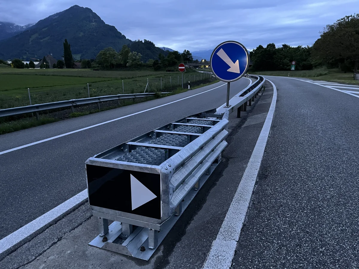 Ein blauweißes Verkehrsschild mit einem Abbiegepfeil steht auf einer Landstraße. Berge und Vegetation sind im Hintergrund sichtbar.