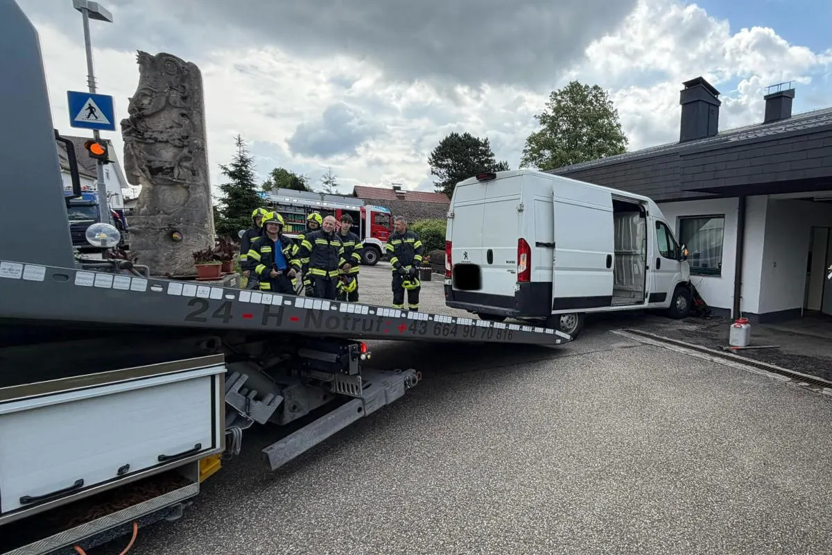 Ein weißer Lieferwagen steht auf einer Abschleppvorrichtung vor einem Gebäude. Im Hintergrund sind mehrere Feuerwehrleute und ein Feuerwehrauto zu sehen.