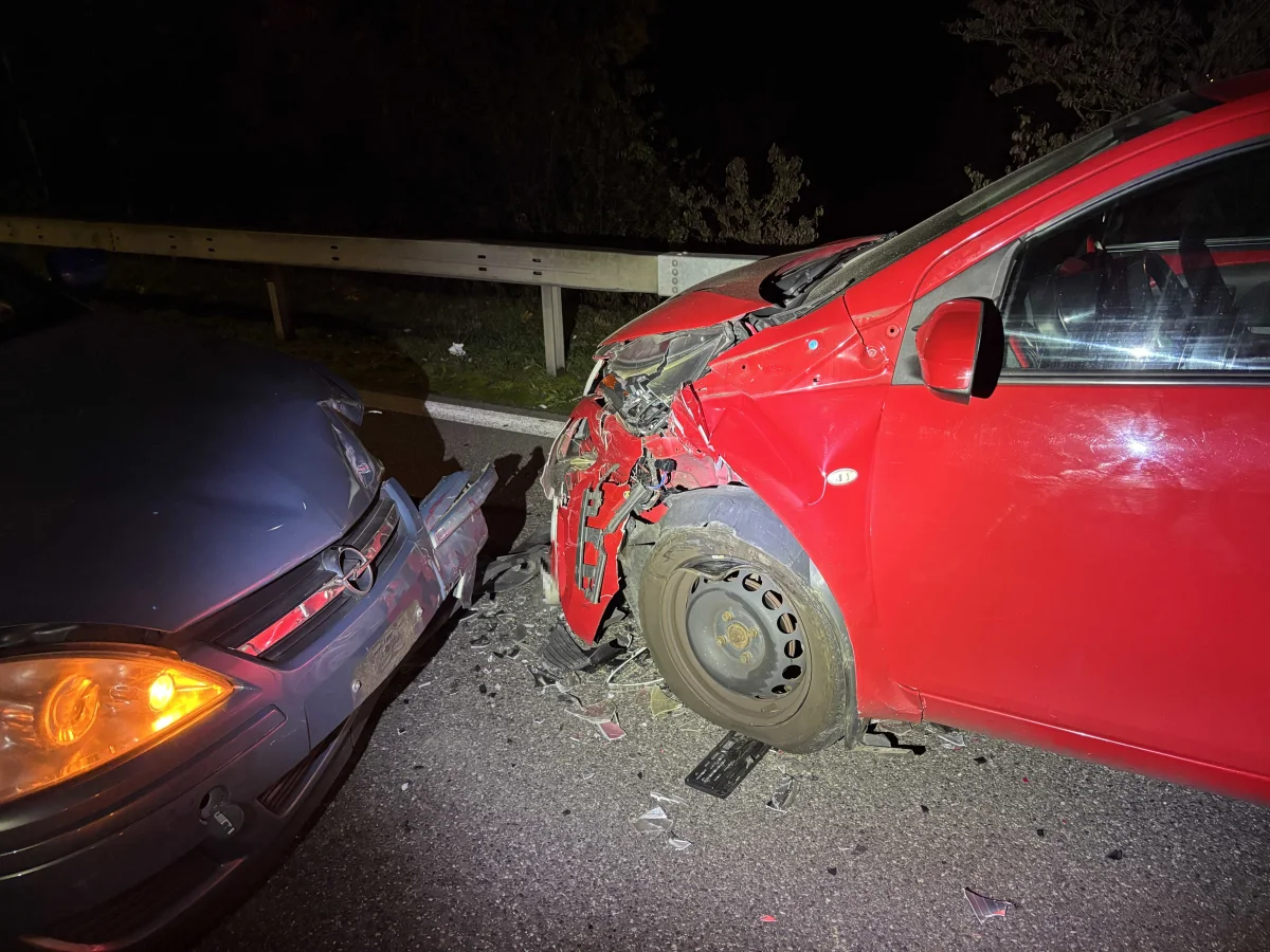 Ein rotes und ein dunkles Auto sind frontal aufeinander geprallt. Die Straße ist dunkel, was auf Nacht hindeutet.