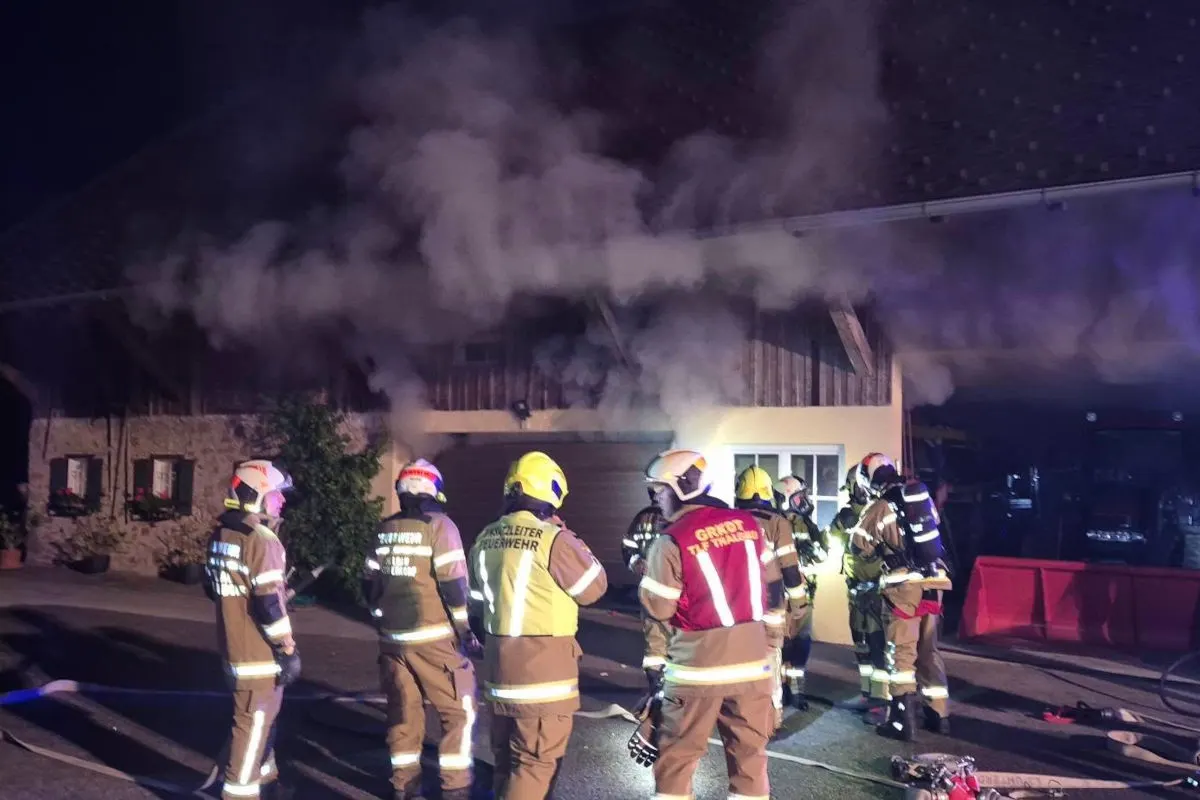 Eine Gruppe von Feuerwehrleuten steht vor einem rauchenden Gebäude in der Nacht. Das Gebäude zeigt deutliche Rauchentwicklung aus Fenstern und Türen.