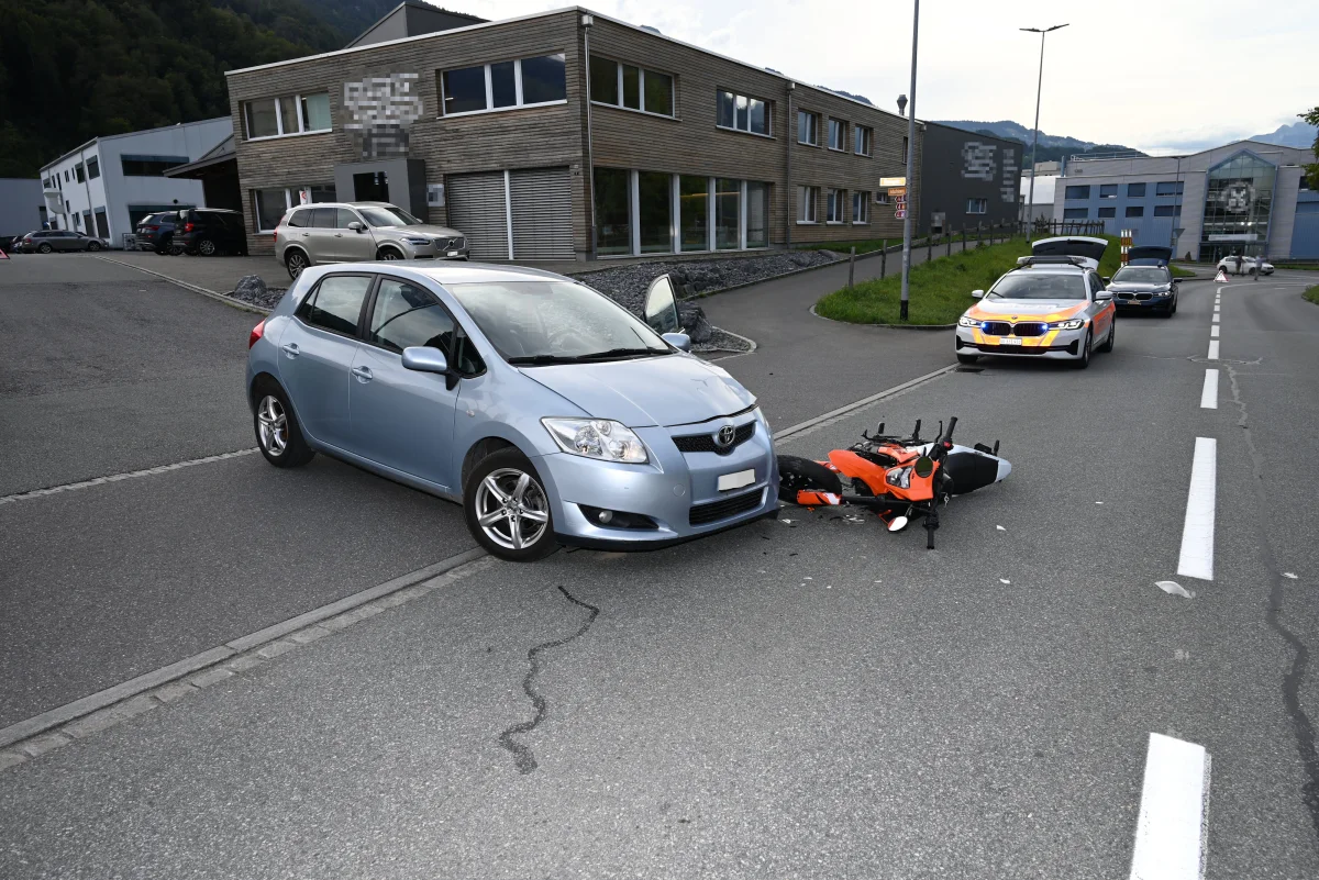 Ein blauer PKW steht mit einem umgestürzten orangefarbenen Motorrad auf der Straße. Ein Polizeifahrzeug ist im Hintergrund zu sehen.
