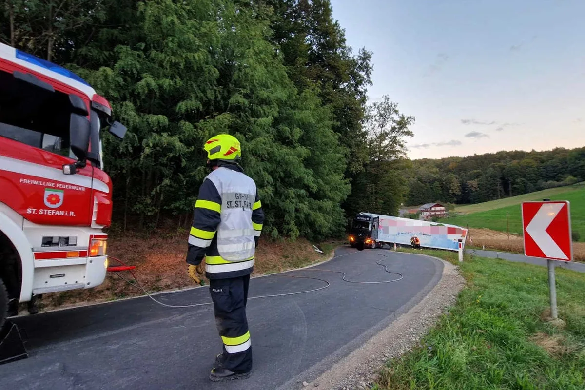 Ein Feuerwehrmann steht auf einer Straße neben einem Feuerwehrfahrzeug. Ein Lastwagen steckt in einer Kurve im Hintergrund fest.