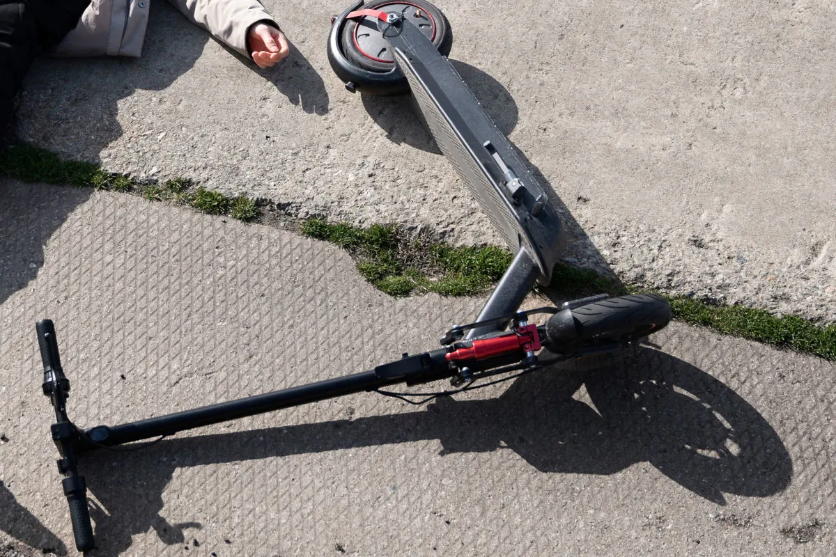 Ein E-Scooter liegt umgestürzt auf einem Gehweg. Eine Person liegt daneben mit einem ausgestreckten Arm.