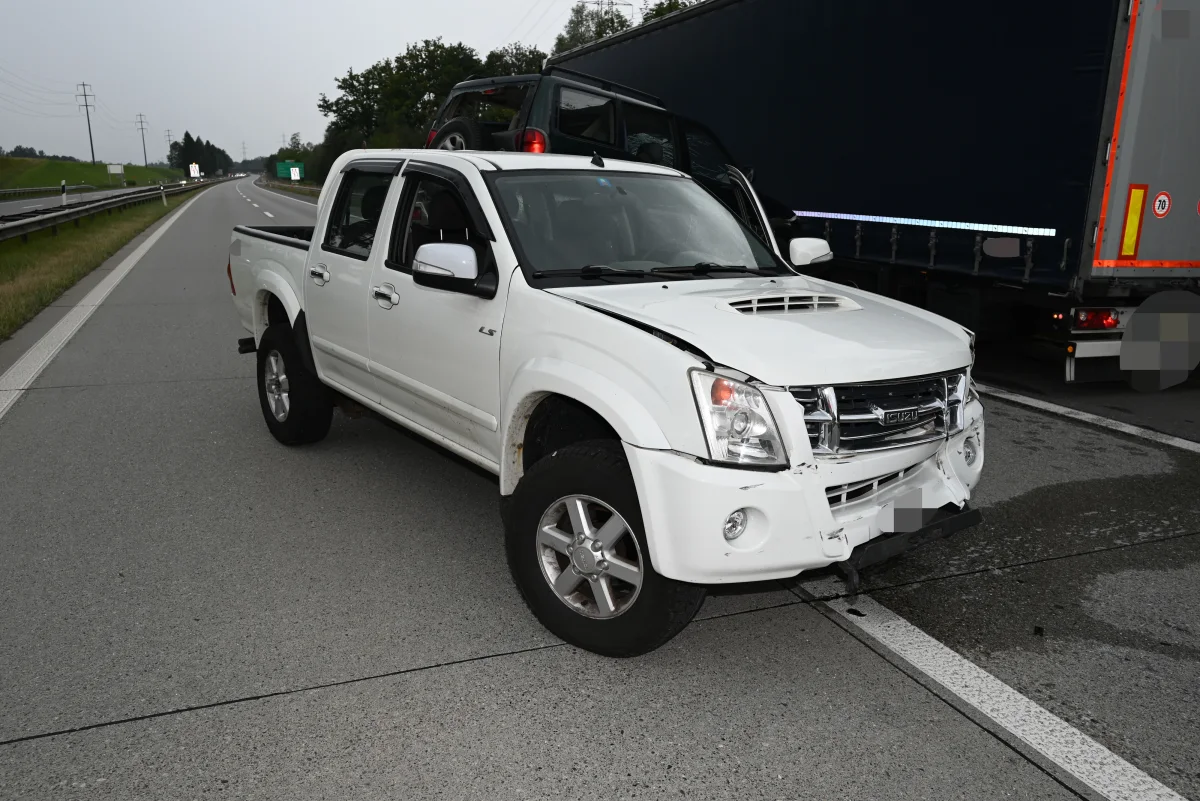 Ein weißer Pickup mit Frontschäden steht auf einer Autobahn. Daneben ist ein dunkler LKW zu sehen.