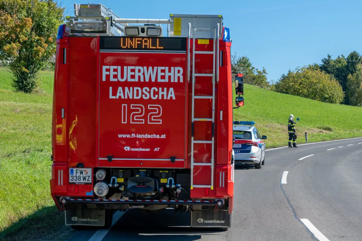 Ein rotes Feuerwehrfahrzeug steht am Straßenrand mit einer digitalen Anzeige 'Unfall'. Im Hintergrund sind ein Polizeiwagen und ein Feuerwehrmann zu sehen.