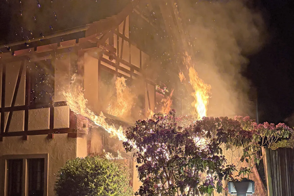 Ein Fachwerkhaus steht nachts in Flammen, mit deutlich sichtbarem Feuer und Rauchentwicklung. Das Feuer hat das Dach erreicht und es sind Glutpartikel in der Luft sichtbar.