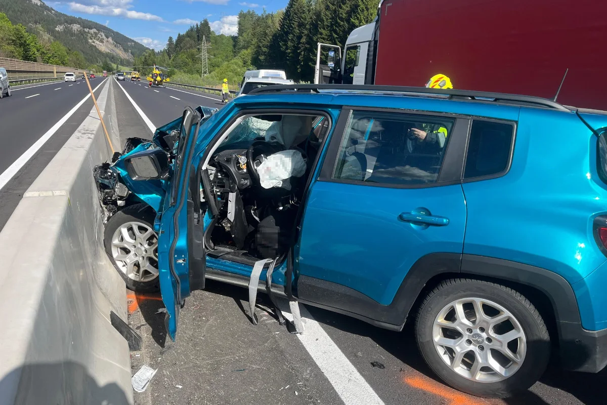 Ein leuchtend blaues Auto steht schwer beschädigt neben einer Betonleitplanke auf einer Autobahn. Es ist ein sonniger Tag mit klarer Sicht.