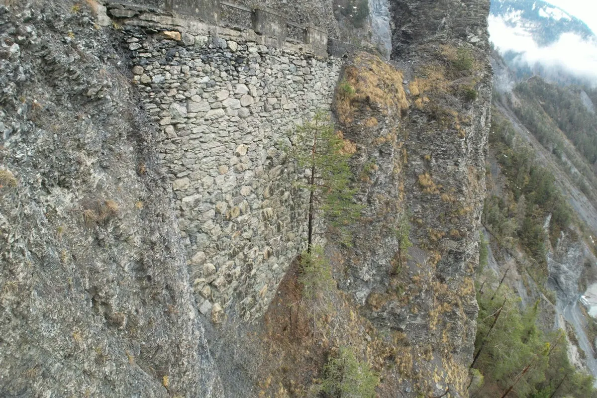 Eine steinerne Stützmauer zieht sich entlang einer felsigen Steilwand, umgeben von Bäumen und alpiner Landschaft.