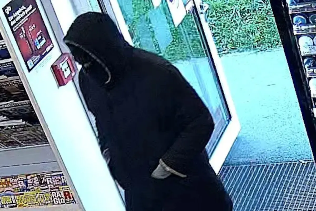Eine vermummte Person in dunkler Kleidung tritt durch die offene Glastür in einen Laden ein. An der Wand ist ein Brandmelder zu sehen.