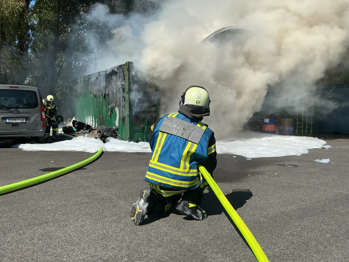 Ein Feuerwehrmann kniet vor einem rauchenden Container und löscht das Feuer mit Schaum. Ein weiterer Feuerwehrmann steht in der Nähe eines Fahrzeugs.